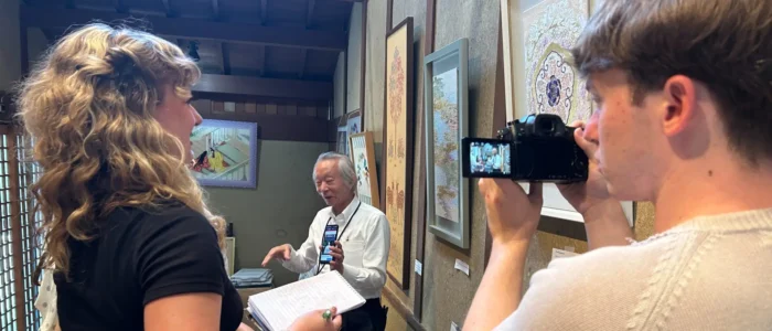 A young woman with a notebook and a young man with a camera interview a Japanese man in a weaving workshop